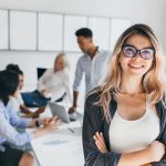 Blonde female executive posing with smile and arms crossed during brainstorm with managers. Indoor portrait of european student spending time in hall with asian and african friends..